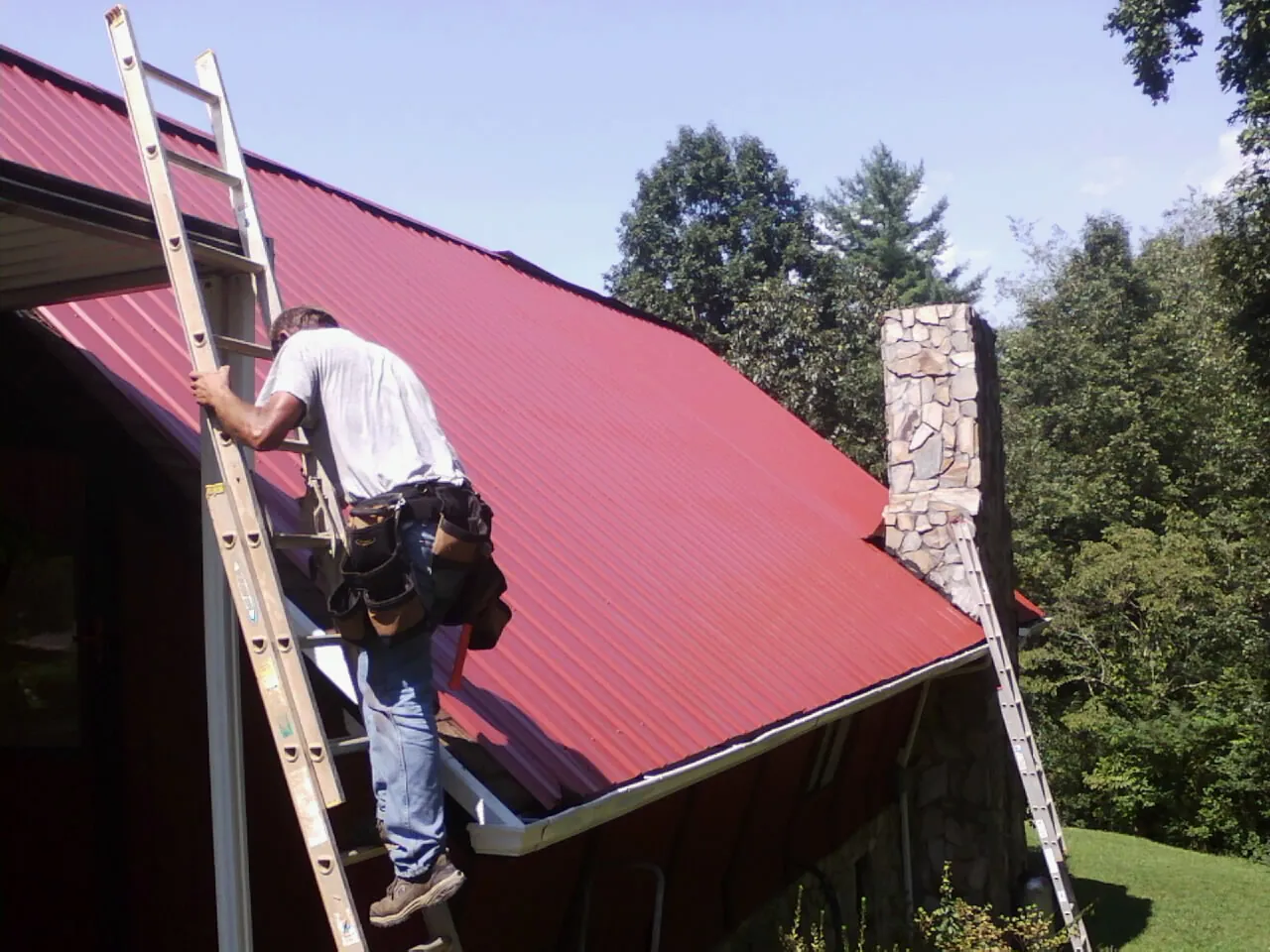 Roofer on a ladder beside a red metal roof and stone chimney — Soffit & Fascia Repair by Roofing Brothers in Bloomfield